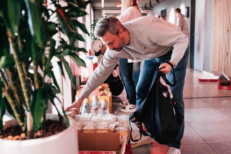 Ein Mann in einem grauen Pullover und Jeans beugt sich vor, um ein Geschenk aus einer Kiste zu nehmen. Er hält eine schwarze Tasche mit einem Fußball-Logo in der Hand. Im Hintergrund sind weitere Personen und eine Pflanze in einem Topf zu sehen.