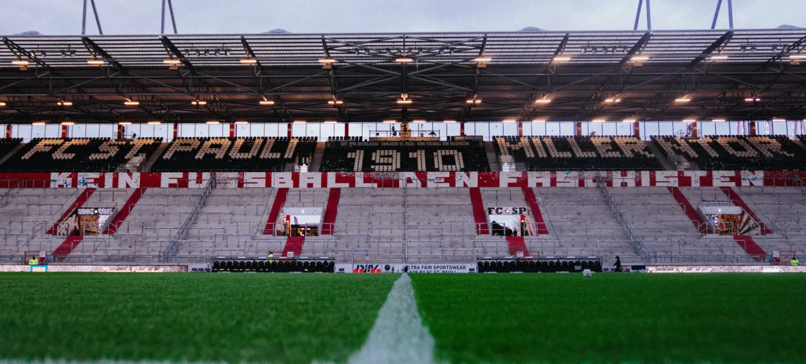 Ein Fußballstadion mit einer großen Choreografie der Fans. Die Tribüne zeigt eine Botschaft in großen Buchstaben, die 'KEIN FUSSBALL DEN FASCHISTEN' lautet. Das Stadion ist teilweise beleuchtet, und die Sitzplätze sind mit roten und schwarzen Elementen gestaltet. Im Vordergrund ist der grüne Rasen des Spielfelds zu sehen.