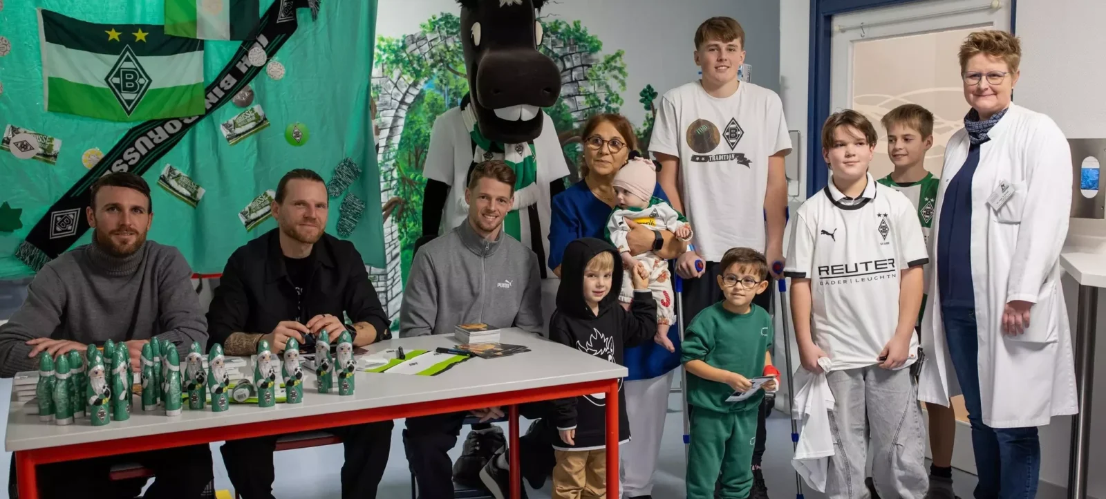Eine Gruppe von Menschen, darunter Kinder und Erwachsene, steht vor einem Tisch mit verschiedenen Produkten. Die Personen tragen Kleidung mit dem Logo des Borussia Mönchengladbach, darunter T-Shirts und Jacken. Im Hintergrund sind eine große Puppe in Form eines Pferdes und eine grüne Wand mit dem Vereinslogo zu sehen. Auf dem Tisch sind mehrere grüne Flaschen und andere Produkte mit dem Vereinslogo angeordnet. Die Atmosphäre wirkt fröhlich und gesellig.