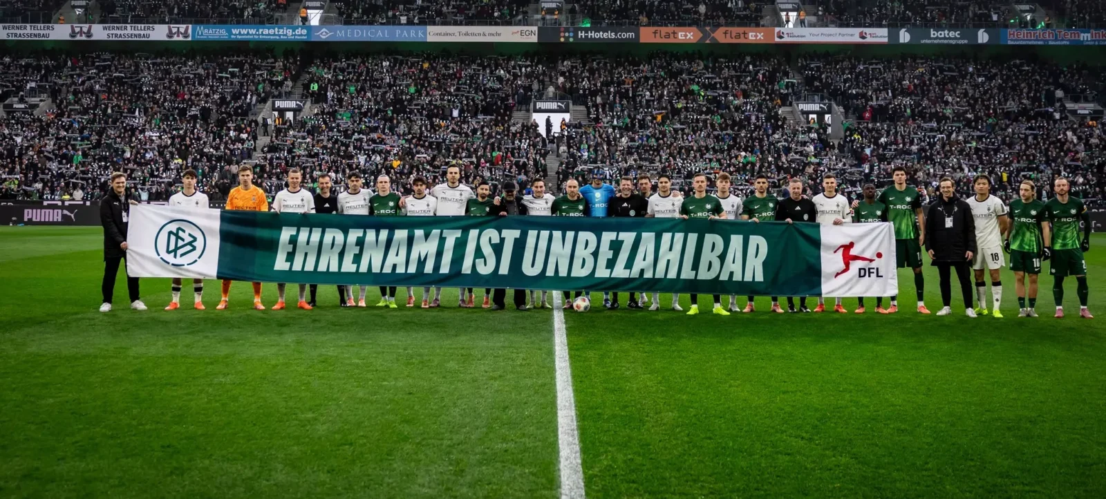 Zwei Fußballmannschaften stehen vor einem Spiel im Stadion. Sie halten ein großes Banner mit der Aufschrift 'Ehrenamt ist unbezahlbar'. Im Hintergrund sind zahlreiche Zuschauer auf den Tribünen zu sehen.