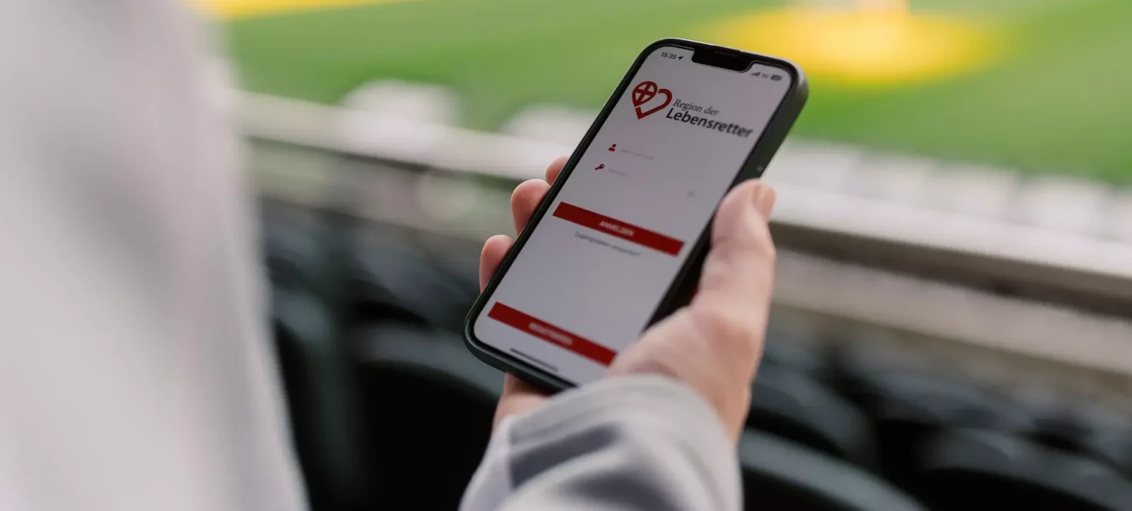 Eine Hand hält ein Smartphone mit der App "Region der Lebensretter" vor einem Fußballfeld.