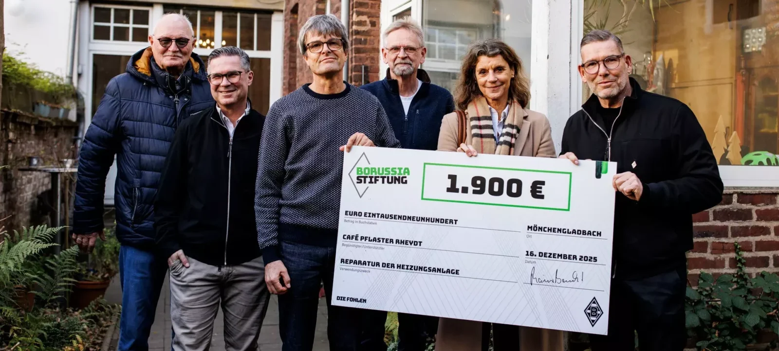 Eine Gruppe von fünf Personen steht vor einem Gebäude und hält einen großen Scheck der Borussia Stiftung im Wert von 1.900 Euro. Der Scheck ist für die Reparatur der Heizungsanlage im Café Pflaster Rheydt in Mönchengladbach bestimmt und datiert auf den 16. Dezember 2025. Die Personen lächeln und wirken zufrieden.