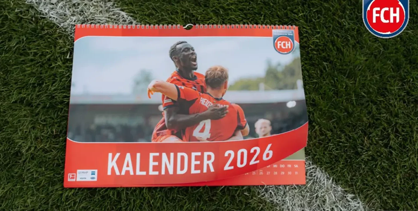 Ein Kalender 2026 des 1. FC Heidenheim liegt auf einem Rasen. Auf dem Kalender sind zwei Fußballspieler in roten Trikots zu sehen, die sich freuen. Im Hintergrund ist das Logo des 1. FC Heidenheim zu erkennen.