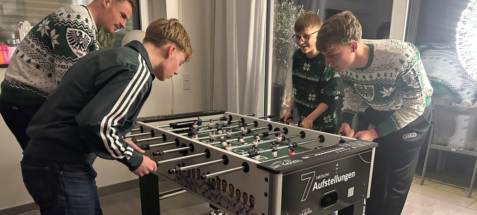 Vier Personen spielen gemeinsam an einem Tischfußball. Die Spieler sind in weihnachtlichen Pullovern gekleidet und scheinen sich in einem festlich dekorierten Raum aufzuhalten.