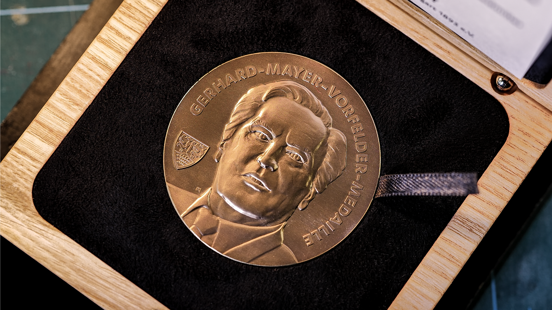 Eine Medaille mit dem Porträt von Gerhard-Mayer-Vorfelder. Die Medaille ist in einem Holzrahmen mit schwarzem Samtuntergrund platziert. Die Inschrift auf der Medaille lautet 'GERHARD-MAYER-VORFELDER' und 'ETTLE' sowie ein kleines Emblem.