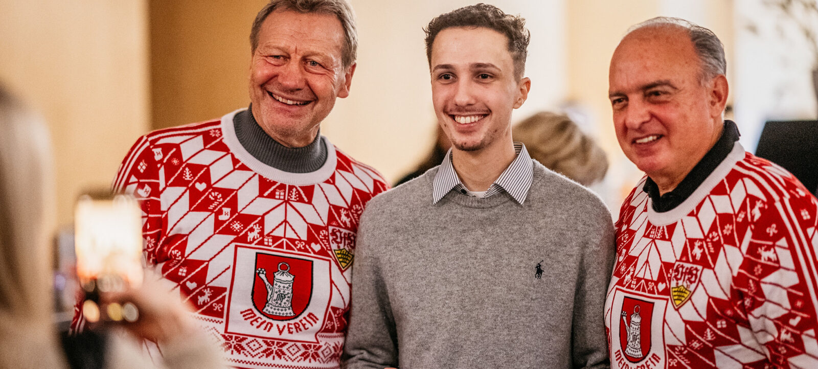 Drei Männer lächeln für ein Selfie, zwei von ihnen tragen Weihnachts-Pullunder mit dem Logo "Mein Verein" und einem Wappen. Der mittlere Mann trägt einen grauen Pullover und hält eine rote Karte mit der Aufschrift "FRITZ!" in der Hand.