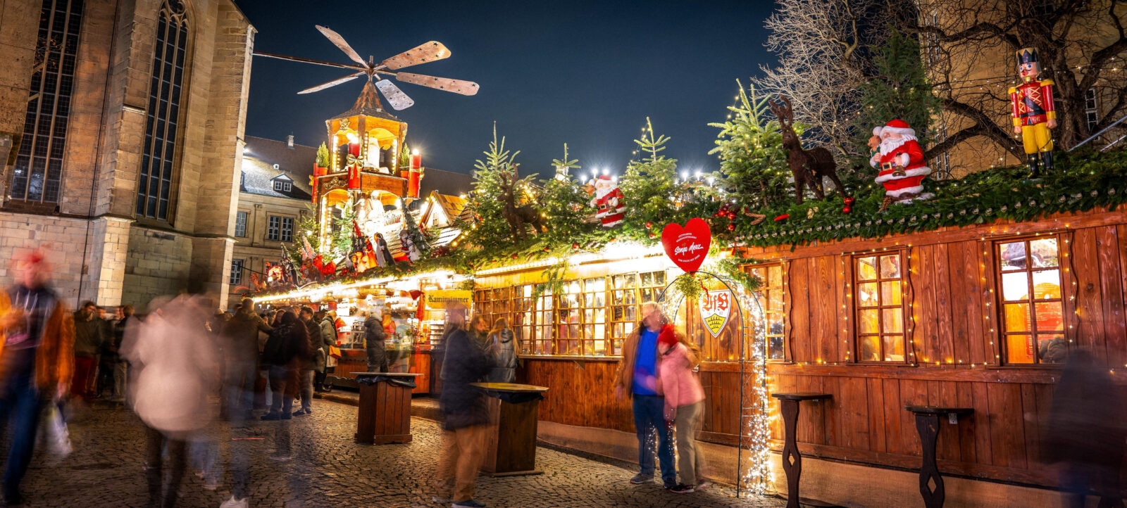 Ein belebter Weihnachtsmarkt bei Nacht mit festlich geschmückten Holzhütten. Die Hütten sind mit Lichtern und Tannenzweigen verziert und haben ein weihnachtliches Dach mit einem großen, sich drehenden Windrad. Menschen sind zu sehen, die sich unterhalten und die Atmosphäre genießen. Im Vordergrund steht ein Schild mit einem Herz und der Aufschrift "Schmeckt nach mehr".