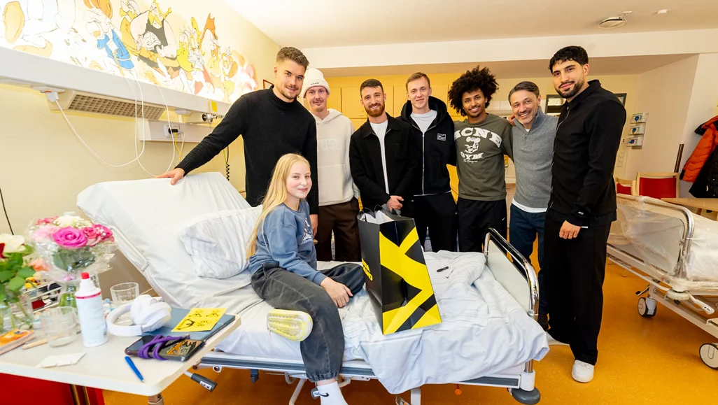 Eine Gruppe von Menschen, darunter Fußballspieler, steht in einem Krankenhauszimmer. Ein junges Mädchen sitzt auf einem Krankenbett und hält einen Tablet-Computer mit einem gelb-schwarzen Design. Auf dem Tisch neben dem Bett befinden sich Blumen, medizinische Utensilien und andere Gegenstände. Die Gruppe lächelt und scheint sich zu freuen.