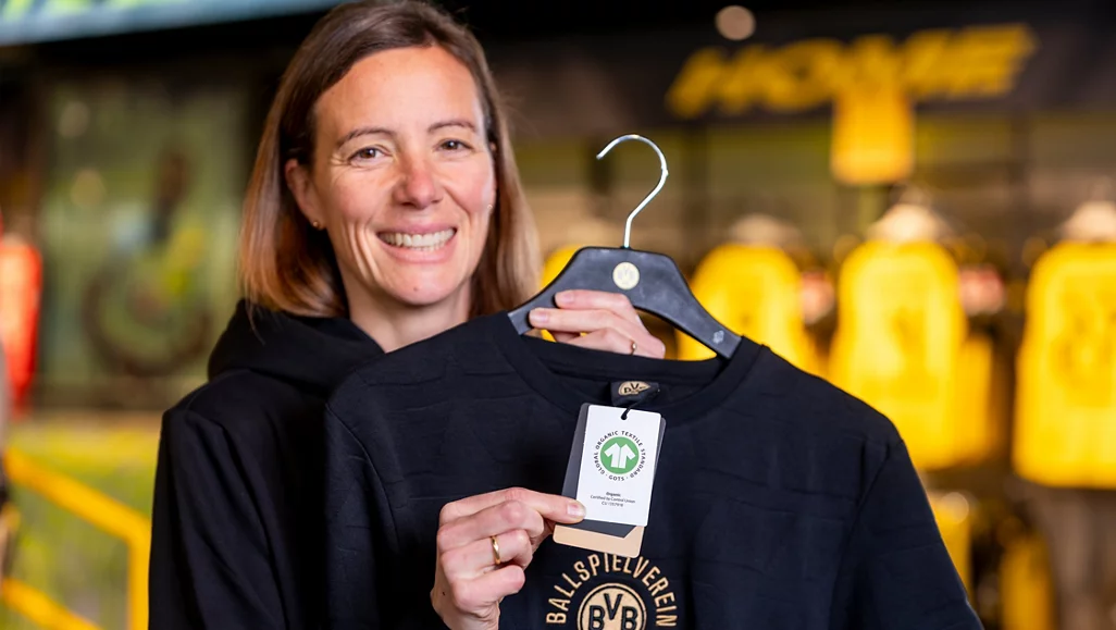 Eine lächelnde Frau hält ein schwarzes T-Shirt mit dem Aufdruck "BALLSPIELVEREIN BVB" hoch. Das T-Shirt hat ein grünes Etikett mit einem Siegel, das auf Nachhaltigkeit hinweist. Im Hintergrund sind gelbe Jacken zu sehen.