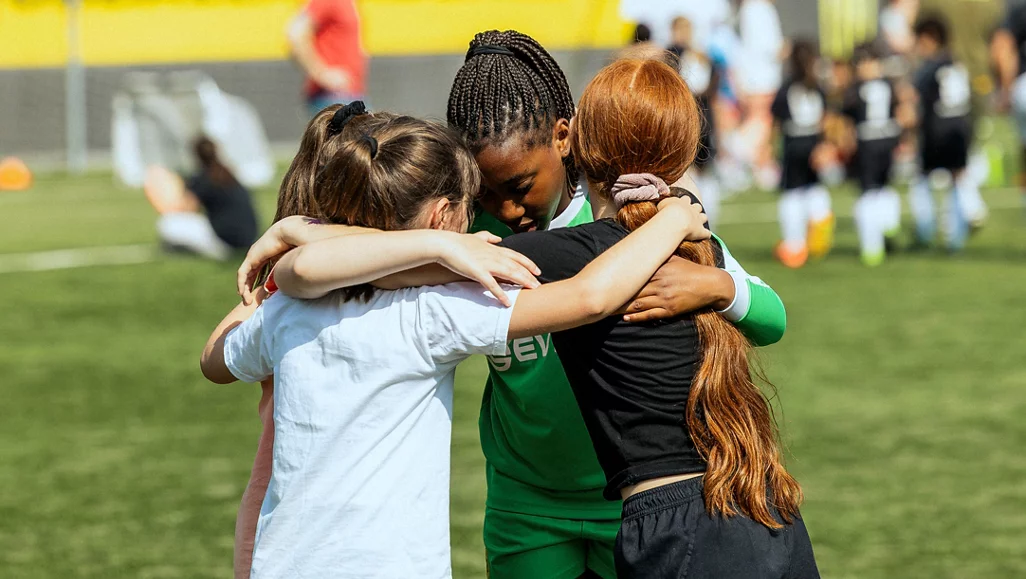 Drei Kinder umarmen sich auf einem Fußballfeld. Sie tragen Sportkleidung und scheinen sich gegenseitig zu unterstützen. Im Hintergrund sind weitere Kinder und Jugendliche zu sehen, die ebenfalls Sportkleidung tragen und sich auf dem Feld aufhalten.