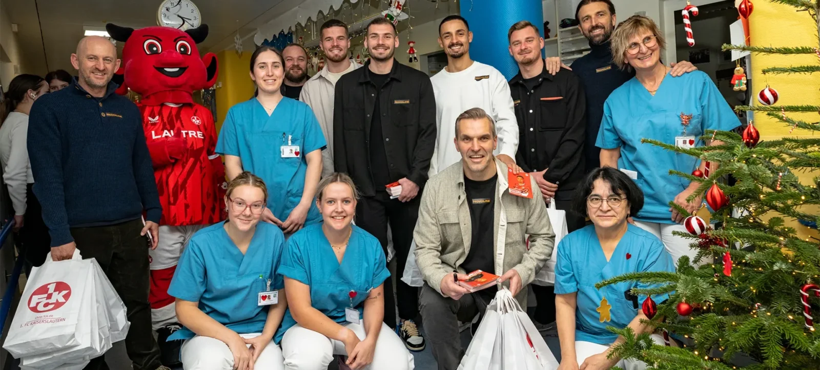Gruppenfoto von Mitarbeitern eines Krankenhauses mit einem Maskottchen und einem Fußballspieler. Die Gruppe steht vor einem weihnachtlich geschmückten Baum und hält Tüten mit Geschenken. Die Mitarbeiter tragen blaue Krankenhauskleidung, während der Fußballspieler und einige andere Personen in Alltagskleidung gekleidet sind.
