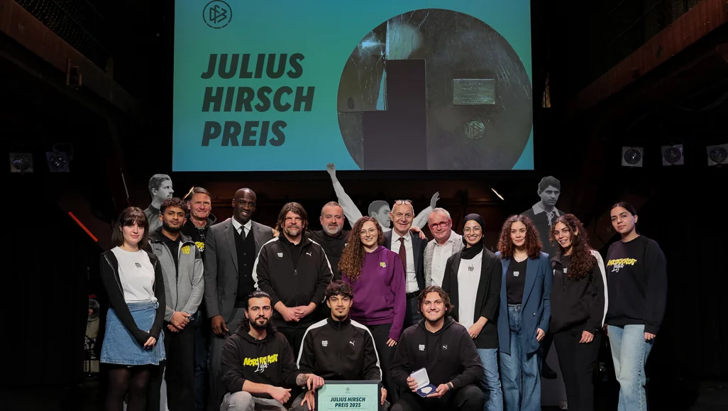 Gruppenfoto der Teilnehmer und Organisatoren des Julius Hirsch Preises. Die Gruppe steht vor einer großen Leinwand mit dem Text 'Julius Hirsch Preis' und einem Bild eines Fußballstadions. Die Personen sind lässig und professionell gekleidet, einige halten Preise in den Händen. Im Vordergrund sitzen drei Personen, darunter ein Preisträger mit einer Urkunde.