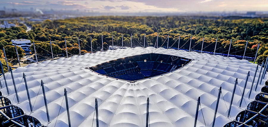 Luftaufnahme eines Fußballstadions mit einem einzigartigen, wellenförmigen Dach aus weißem Material, das von zahlreichen Stützen getragen wird. Das Stadion ist von einem ausgedehnten Waldgebiet umgeben, was die Integration von Natur und Sportarchitektur zeigt.