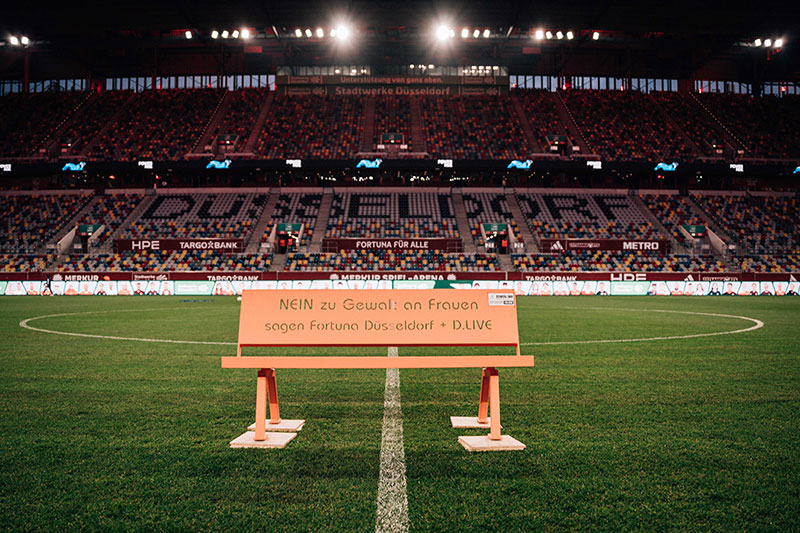Ein Fußballstadion bei Nacht mit einer großen Anzahl von Zuschauern. Im Vordergrund steht ein Schild mit der Aufschrift "NEIN zu Gewalt an Frauen sagen Fortuna Düsseldorf + D.LIVE" auf dem Spielfeld.
