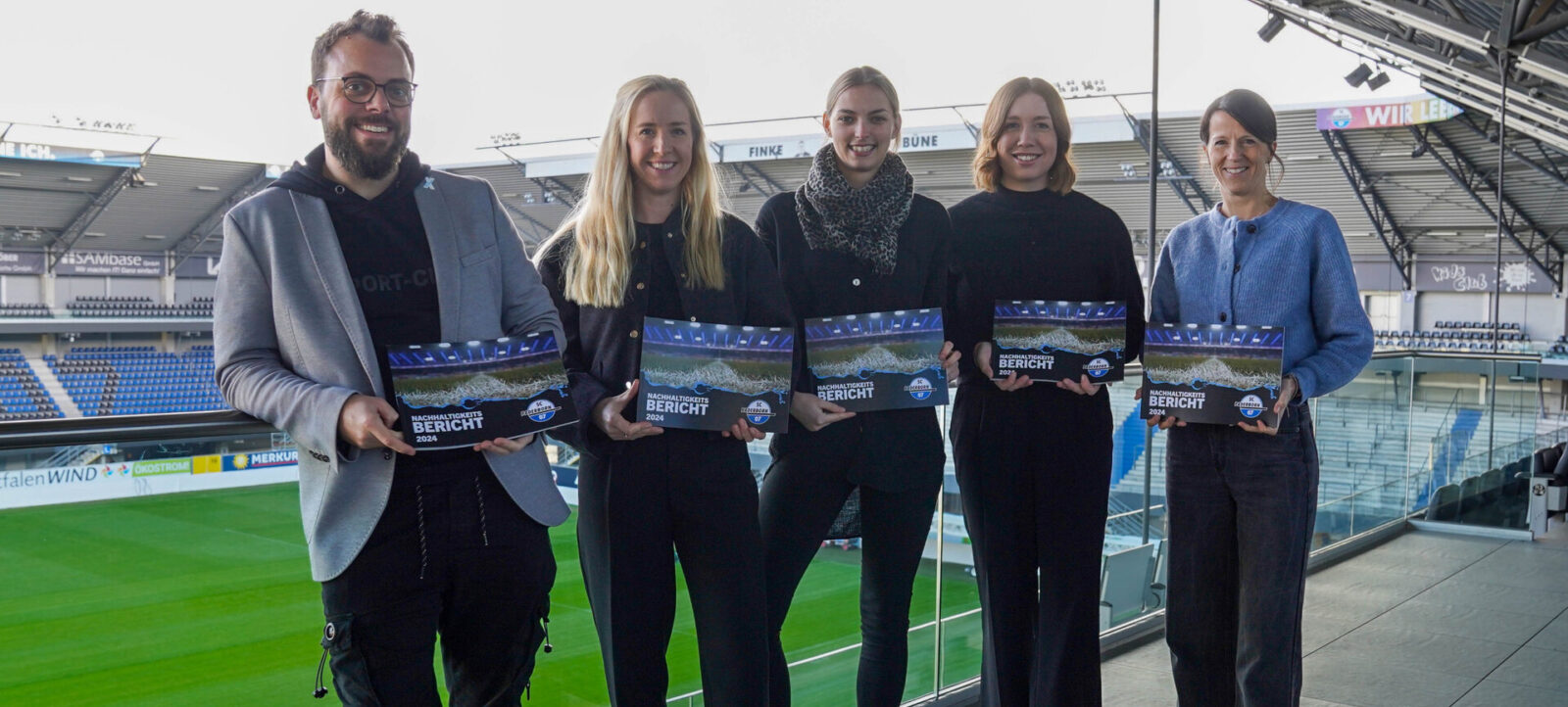 Fünf Personen stehen auf einer Tribüne eines Fußballstadions und halten jeweils ein Exemplar des "Nachhaltigkeitsberichts 2024" in den Händen. Sie lächeln in die Kamera. Im Hintergrund sind Sitzplätze und Werbebanner im Stadion zu sehen.