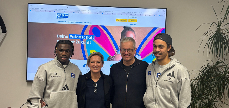 Vier Personen stehen vor einem großen Bildschirm, auf dem eine Website mit dem Logo von PLAN International und dem Text "Deine Patenschaft. Gestalte Zukunft" zu sehen ist. Die Personen tragen Hoodies mit dem Logo des Hamburger SV. Links steht ein junger Mann mit Dreadlocks, daneben eine Frau mit blonden Haaren, gefolgt von einem älteren Mann mit Brille und einem weiteren jungen Mann mit Dreadlocks und einer schwarzen Mütze. Im Hintergrund befindet sich eine Pflanze.