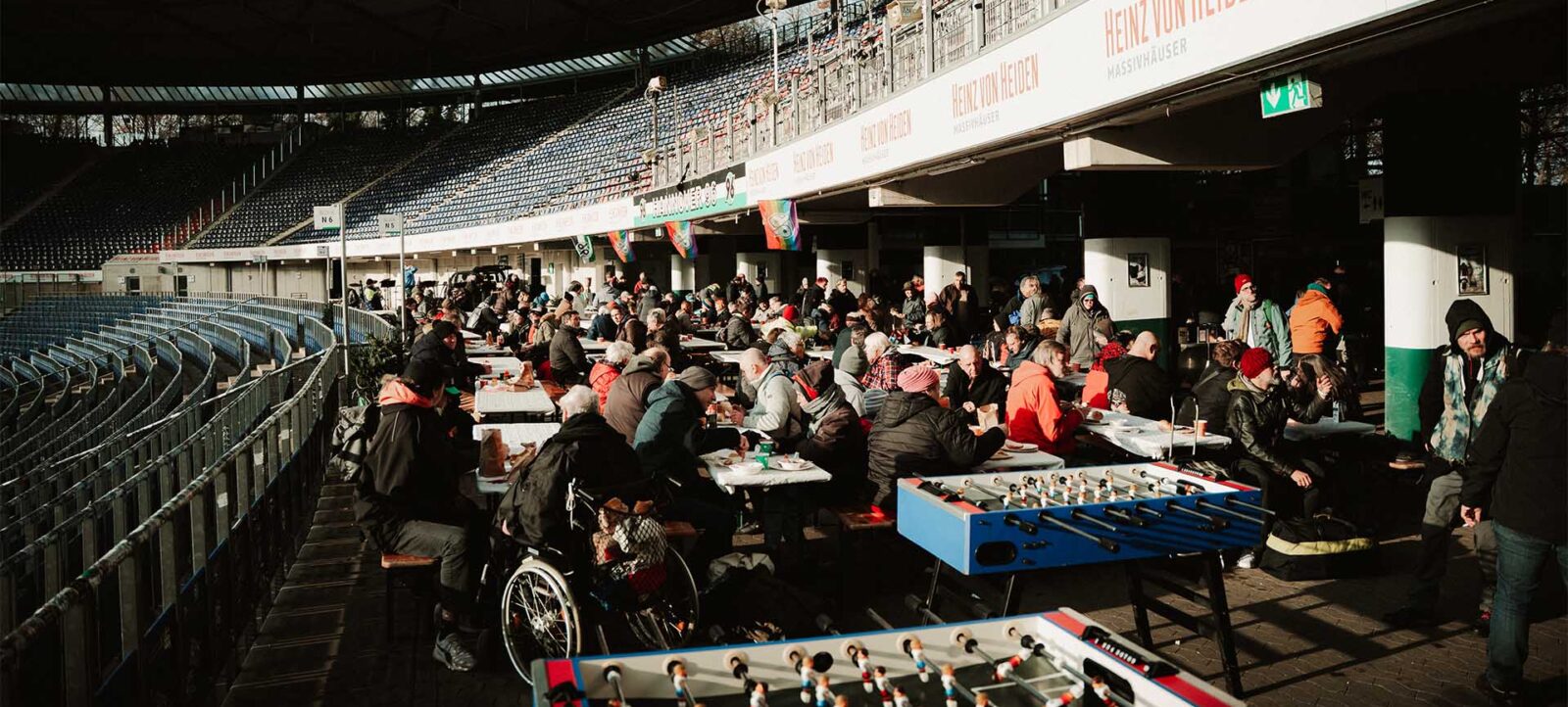 Menschen sitzen an Tischen in einem überdachten Bereich eines Stadions und genießen ein Winterfest. Im Vordergrund sind zwei Kickertische zu sehen. Einige Personen sind in Rollstühlen, und die Atmosphäre wirkt gesellig und gemütlich.