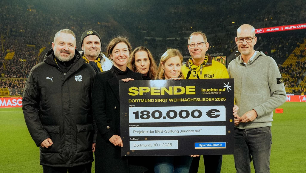 Eine Gruppe von sechs Personen steht auf einem Fußballfeld und hält ein großes Scheckblatt. Das Scheckblatt zeigt eine Spende von 180.000 Euro für das Projekt "Dortmund singt Weihnachtslieder 2025" der BVB-Stiftung "leuchte auf". Die Personen lächeln und posieren für das Foto. Im Hintergrund sind Zuschauer auf den Tribünen zu sehen.