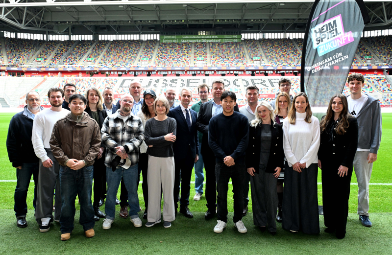 Eine Gruppe von Menschen steht auf dem Rasen eines Stadions und lächelt in die Kamera. Im Hintergrund sind zahlreiche Zuschauer auf den Tribünen zu sehen. Ein Banner mit der Aufschrift "Gemeinsam für Deine Chancen" ist sichtbar.