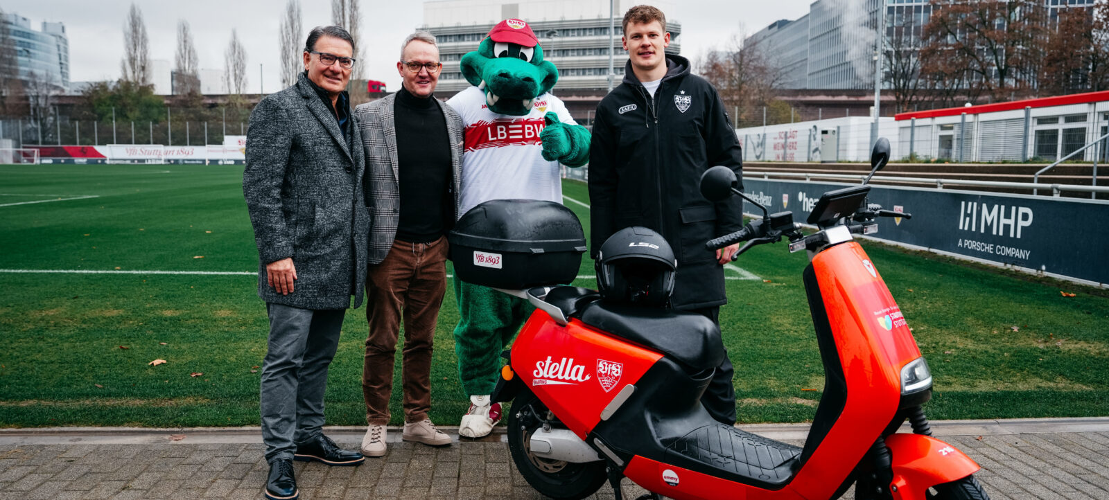 Vier Personen stehen auf einem Fußballfeld neben einem roten E-Roller. Zwei Männer in Anzug und Pullover, eine Person im Maskottchen-Kostüm und ein junger Mann in Jacke und Jeans. Der Roller ist mit einem Helm und einem Rucksack ausgestattet und trägt das Logo von Stella Artois und dem VfB Stuttgart. Im Hintergrund sind Tribünen und moderne Gebäude zu sehen.