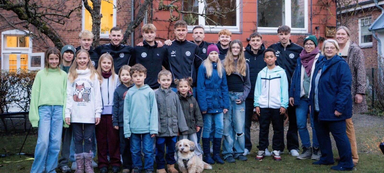 Eine Gruppe von Kindern, Jugendlichen und Erwachsenen steht vor einem roten Backsteingebäude und lächelt in die Kamera. Die Kinder und Jugendlichen tragen verschiedene warme Kleidung, darunter Pullover, Jacken und Mützen. Einige Erwachsene sind ebenfalls anwesend und tragen ebenfalls warme Kleidung. Ein kleiner Hund sitzt vorne in der Gruppe. Die Atmosphäre wirkt fröhlich und gesellig.