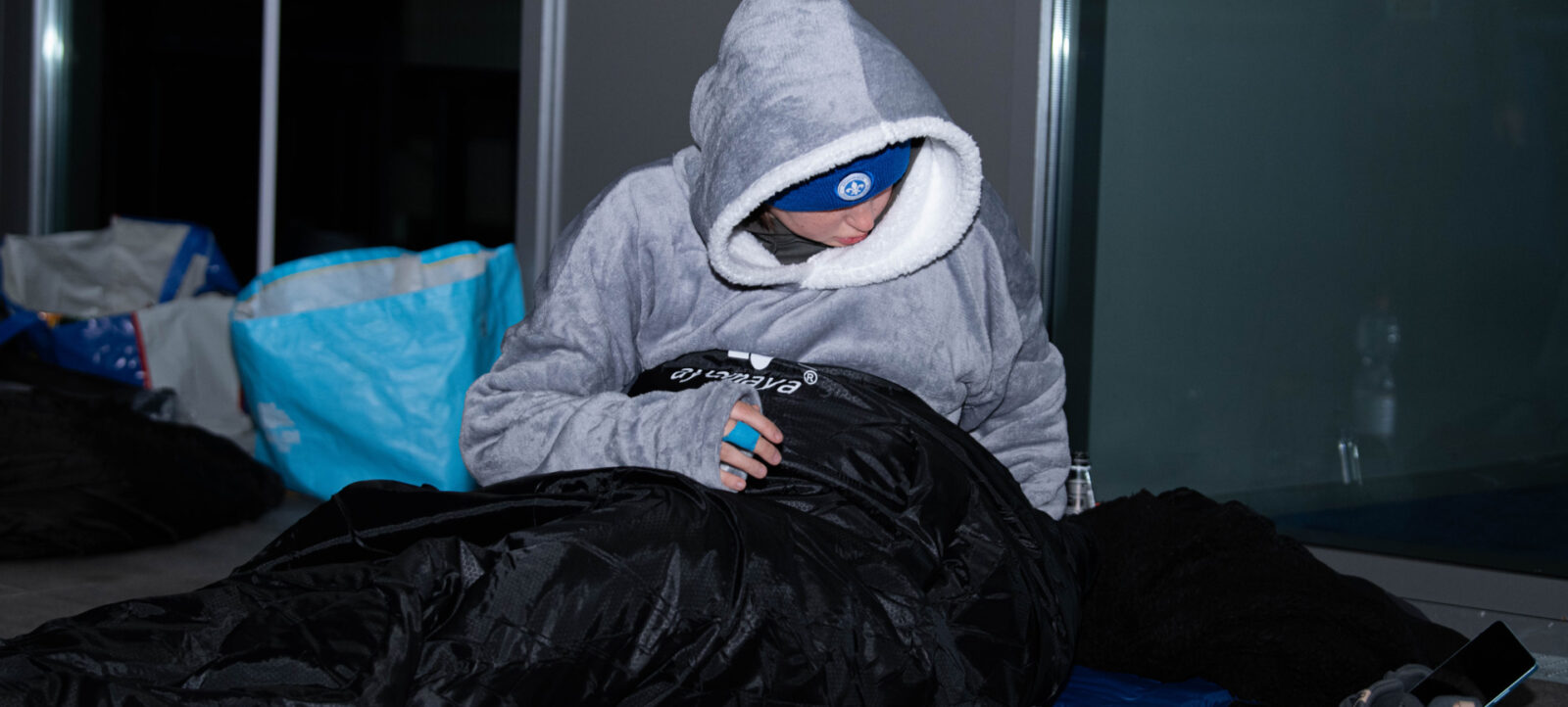 Eine Person schläft in einem Schlafsack auf dem Boden, eingekuschelt in eine graue Kapuzenjacke und mit einer Schlafmaske. Neben der Person liegt eine blaue Isomatte und ein Smartphone auf dem Boden. Im Hintergrund sind ein blauer Sack und eine Glaswand zu sehen.