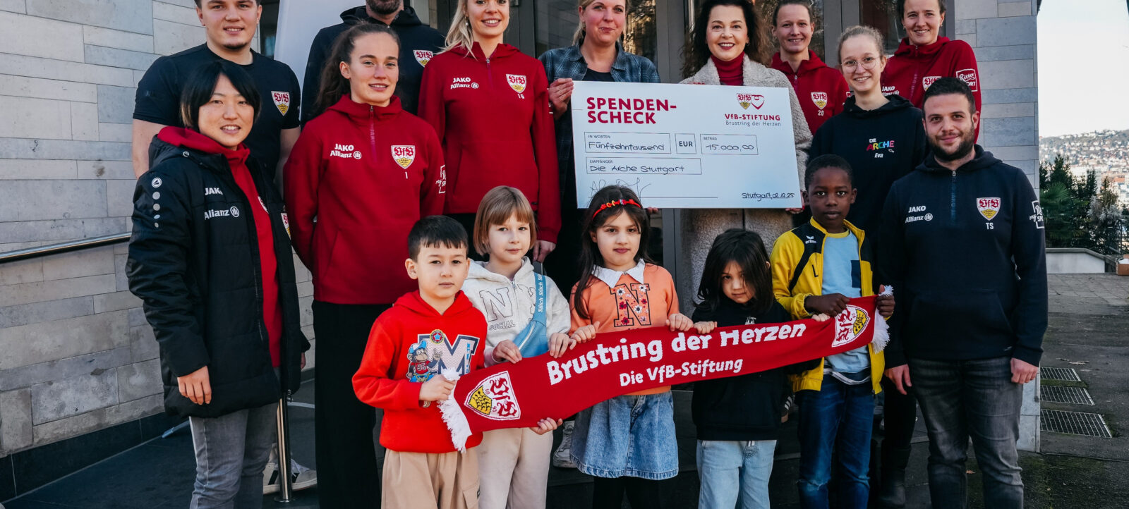 Eine Gruppe von Erwachsenen und Kindern hält ein großes Scheck über 15.000 Euro für die "Die Arche Stuttgart" von der VfB-Stiftung. Die Erwachsenen tragen Jacken mit dem Logo des VfB Stuttgart und dem Sponsor JAKO. Die Kinder halten ein rotes Banner mit der Aufschrift "Brustring der Herzen Die VfB-Stiftung". Die Gruppe steht vor einem Gebäude und lächelt in die Kamera.