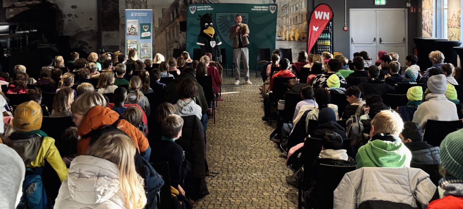 Eine Gruppe von Kindern und Erwachsenen sitzt in einem Raum und hört aufmerksam einer Person zu, die an einem Pult steht und aus einem Buch vorliest. Im Hintergrund ist eine große Maske eines Vogels zu sehen, die Teil einer Wanddekoration ist. Der Raum hat eine rustikale Atmosphäre mit Steinwänden und einem gepflasterten Boden.