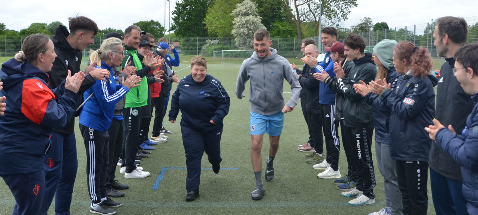 Eine Gruppe von Menschen steht auf einem Fußballfeld und klatscht begeistert, während zwei Personen durch die Mitte gehen und lächeln. Die Teilnehmer tragen verschiedene Jacken und Hoodies, einige mit Logos von Fußballclubs. Im Hintergrund sind weitere Personen und Bäume zu sehen.