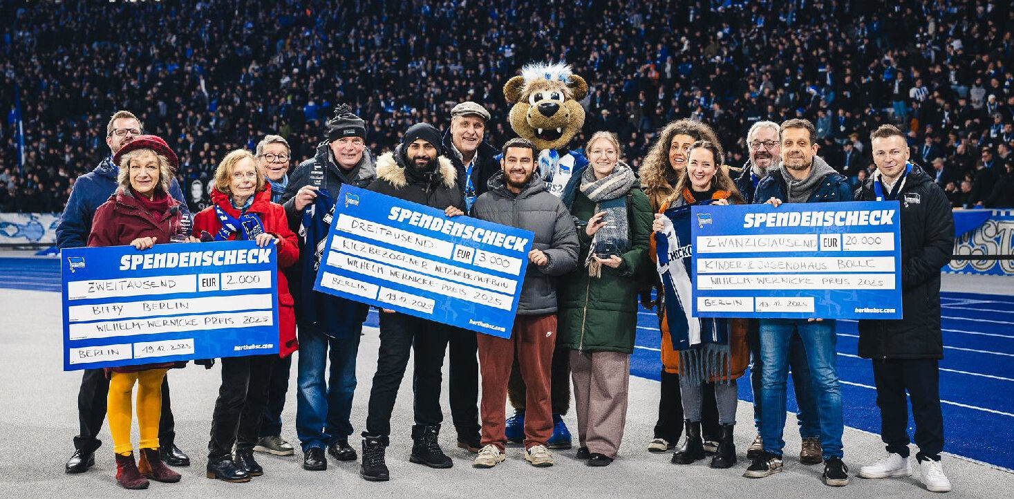 Eine Gruppe von Menschen hält große Schecks mit Spendenbeträgen in den Händen. Sie stehen auf einem Fußballfeld vor einer großen Zuschauermenge. Ein Maskottchen in einem Bärenkostüm ist ebenfalls Teil der Gruppe. Die Schecks sind auf verschiedene Beträge ausgestellt, darunter 2.000 Euro, 3.000 Euro und 20.000 Euro, und sind für verschiedene Projekte bestimmt, wie z.B. "Kiez & Begegnungsstätte Berlin" und "Kinder- & Jugendhaus Bollhagen". Die Veranstaltung findet im Rahmen des Wilhelm-Wernicke-Preises 2025 statt.