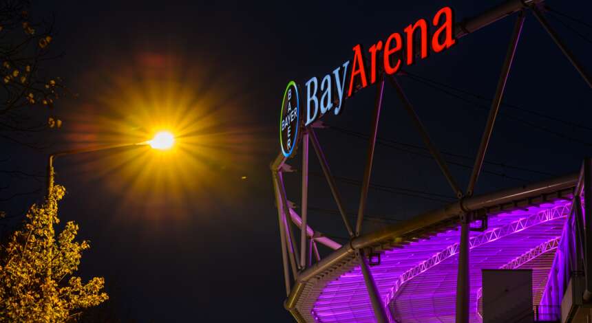 Nachtszene mit der beleuchteten BayArena, die in lila Licht getaucht ist. Das Stadionlogo und der Name 'BayArena' sind deutlich sichtbar. Im Hintergrund ist ein beleuchteter Baum und eine Straßenlaterne zu sehen.
