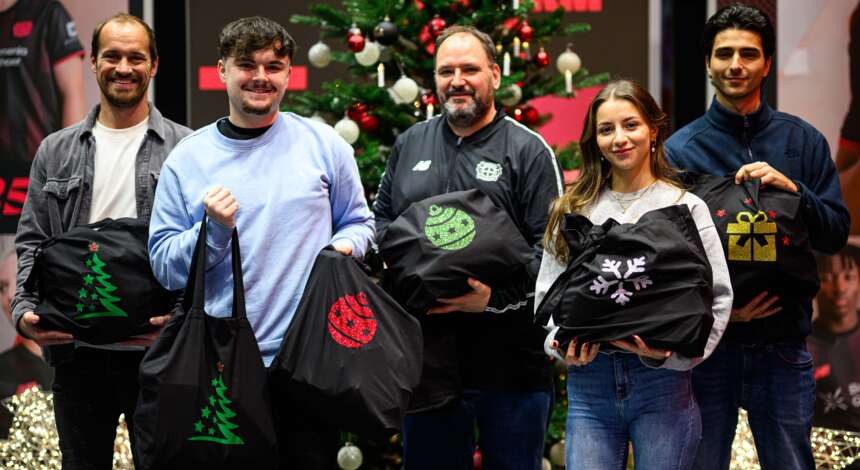 Gruppe von Menschen hält schwarze Stoffbeutel mit Weihnachtsmotiven vor einem geschmückten Weihnachtsbaum. Die Beutel sind mit verschiedenen Weihnachtsdesigns bedruckt, darunter Tannenbäume, Schneeflocken und Geschenke. Die Personen lächeln und stehen nebeneinander, möglicherweise bei einer Weihnachtsfeier oder einem ähnlichen Event.