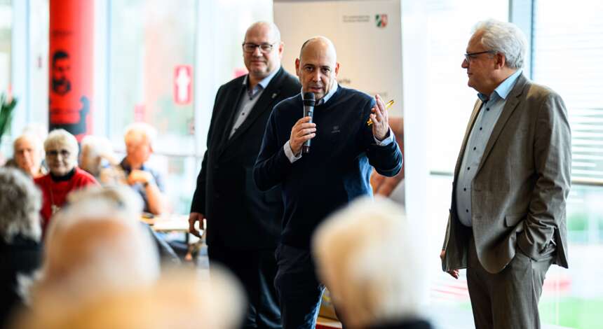 Drei Männer in Anzügen stehen vor einer Gruppe von Menschen. Der Mann in der Mitte hält ein Mikrofon und spricht, während die anderen beiden Männer zuhören. Die Szene findet in einem gut beleuchteten Raum statt, möglicherweise bei einer Veranstaltung oder einem Meeting. Im Hintergrund sind einige Personen zu sehen, die an Tischen sitzen.