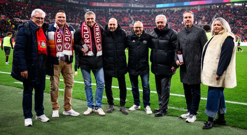 Eine Gruppe von sieben Personen steht auf einem Fußballfeld und lächelt in die Kamera. Sie tragen verschiedene Jacken und Schals, einige mit dem Logo des 1. FC Köln. Im Hintergrund sind viele Zuschauer auf den Tribünen zu sehen.