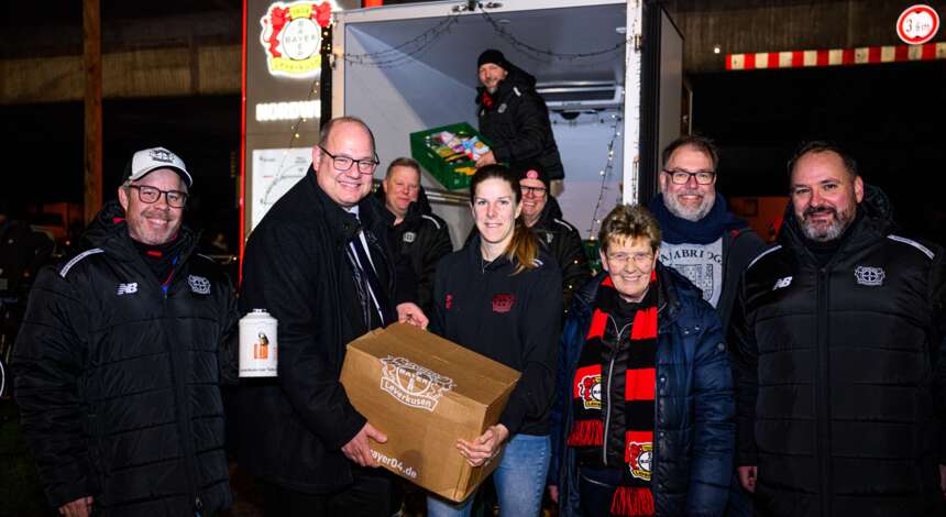 Eine Gruppe von Menschen steht vor einem Transporter und hält eine große Kiste mit dem Logo des 1. FC Köln. Sie lächeln und scheinen sich über eine erfolgreiche Spendenübergabe zu freuen. Die Personen tragen Jacken und einige haben Schals mit den Vereinsfarben des 1. FC Köln. Im Hintergrund ist ein Logo des 1. FC Köln an der Wand des Transporters zu sehen.