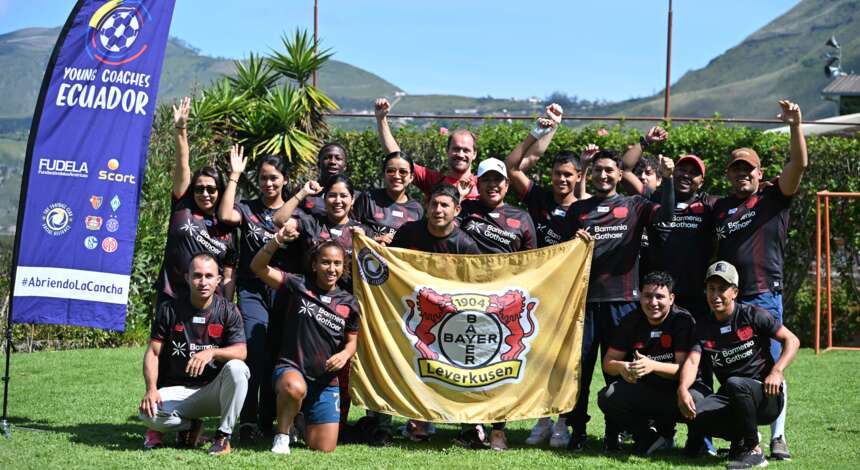 Eine Gruppe von Menschen, darunter Trainer und Spieler, posieren auf einem Fußballfeld und halten ein Banner mit dem Logo von Bayer 04 Leverkusen. Sie tragen T-Shirts mit dem Aufdruck "Bayer 04 Leverkusen" und "Young Coaches Ecuador". Im Hintergrund ist eine Berglandschaft zu sehen, und eine Fahne mit der Aufschrift "Young Coaches Ecuador" ist sichtbar.