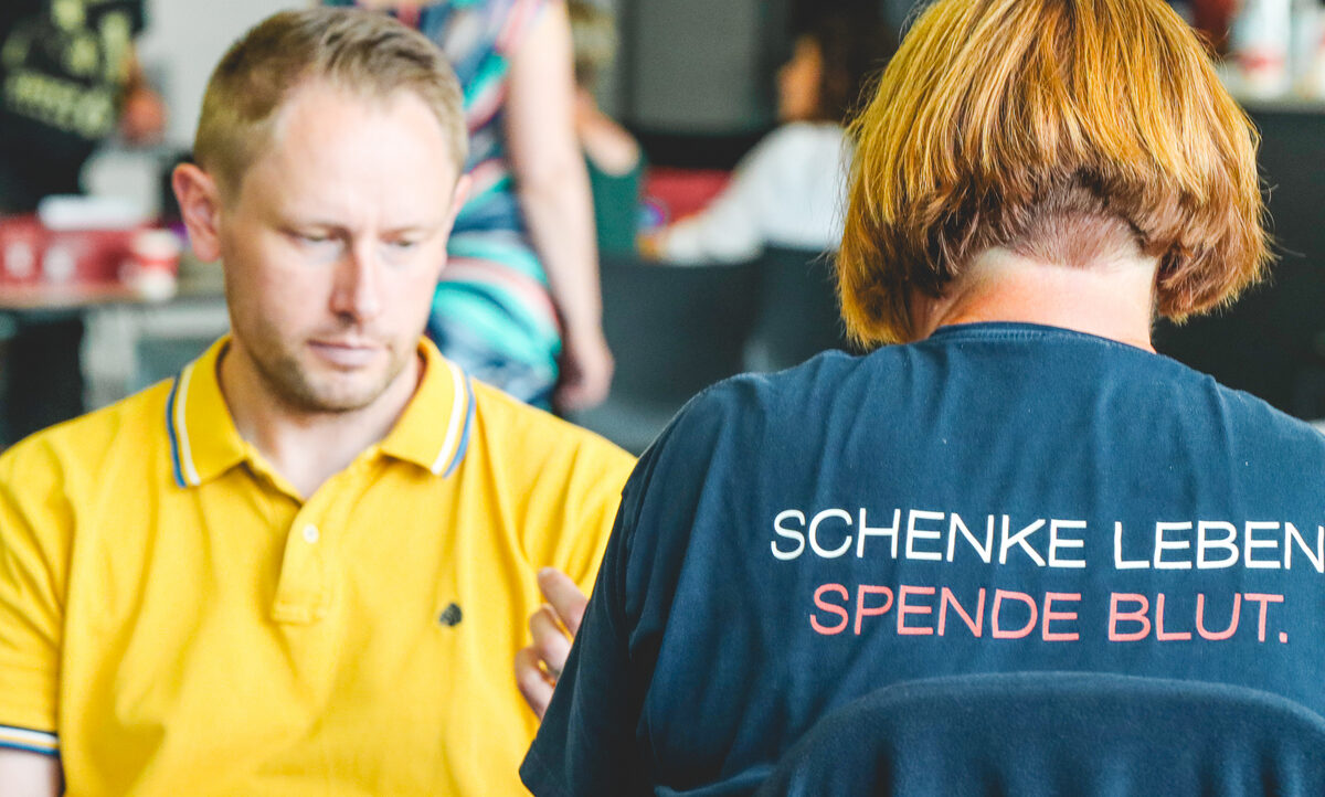 Zwei Männer, einer in einem gelben Hemd und der andere in einem blauen Hemd mit der Aufschrift "Schenke Leben, spende Blut", scheinen sich auf eine Blutspendeaktion vorzubereiten.