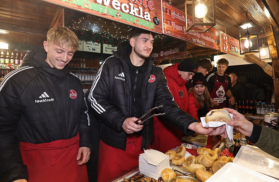 Zwei Personen in schwarzen Jacken mit roten Schürzen stehen an einem Imbissstand und bedienen Kunden. Sie halten jeweils ein Brötchen in der Hand und lächeln. Der Stand ist mit verschiedenen Lebensmitteln und Getränken dekoriert, darunter mehrere Brötchen und Getränkedosen. Im Hintergrund sind weitere Personen zu sehen, die auf das Essen warten.