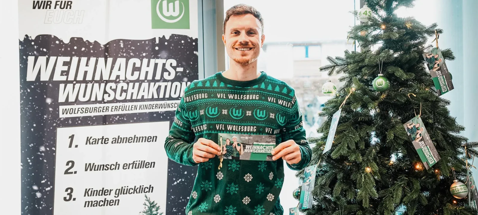 Ein Mann in einem grünen Weihnachts-Pullover mit dem Logo des VFL Wolfsburg hält eine Weihnachtskarte in der Hand. Er steht vor einem Plakat, das die Weihnachtswunschaktion des VFL Wolfsburg beschreibt, bei der Kinderwünsche erfüllt werden. Im Hintergrund ist ein geschmückter Weihnachtsbaum mit grünen und silbernen Kugeln und Bildern von Spielern des VFL Wolfsburg zu sehen.