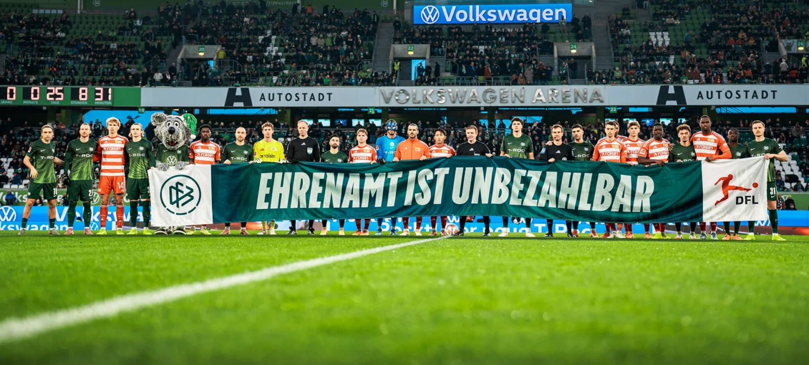 Fußballmannschaft und Ehrenamtliche halten ein Transparent mit der Aufschrift 'Ehrenamt ist unbezahlbar' in der Volkswagen Arena. Die Spieler und Ehrenamtliche stehen vor den Zuschauern und halten das Transparent, das die Logos des VfL Wolfsburg und der DFL zeigt. Im Hintergrund sind die Tribünen mit vielen Zuschauern zu sehen.