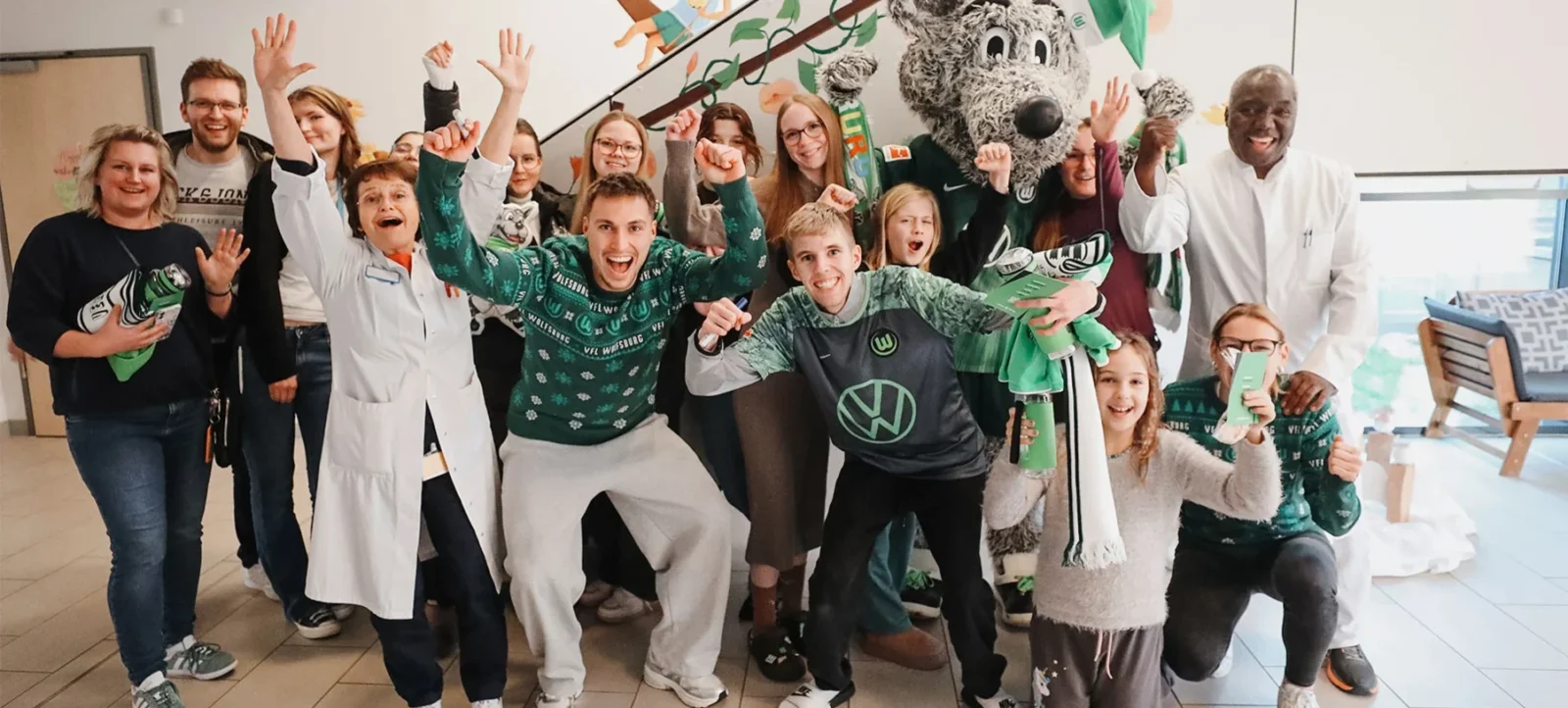 Eine fröhliche Gruppe von Menschen, darunter Kinder, Erwachsene und ein Maskottchen, posiert in einem Krankenhausflur. Sie tragen grüne und weiße Kleidung mit dem Logo des VFL Wolfsburg und halten grüne Luftballons in den Händen. Im Hintergrund ist ein Schild mit der Aufschrift "Willkommen" zu sehen.