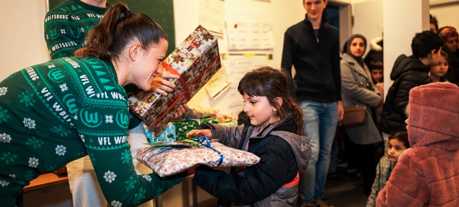Eine Person in einem grünen VFL Wolfsburg Sweatshirt überreicht einem Kind ein Geschenk. Im Hintergrund stehen weitere Personen, darunter einige Kinder, in einem Raum mit einem Tisch voller Geschenke.