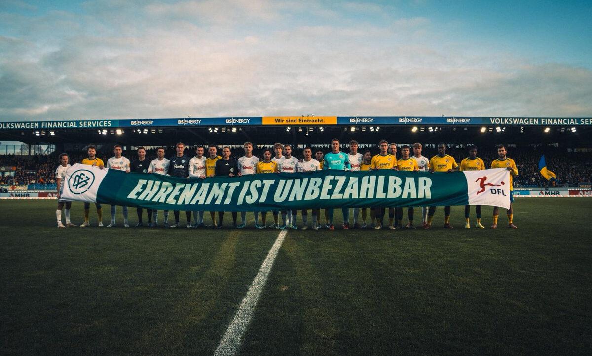 Zwei Fußballmannschaften stehen auf dem Spielfeld und halten ein großes grünes Banner mit der Aufschrift 'Ehrenamt ist unbezahlbar'. Die Spieler tragen Trikots in den Farben Gelb-Schwarz und Weiß. Im Hintergrund sind die Stadiontribünen mit Zuschauern und Werbebanden zu sehen.