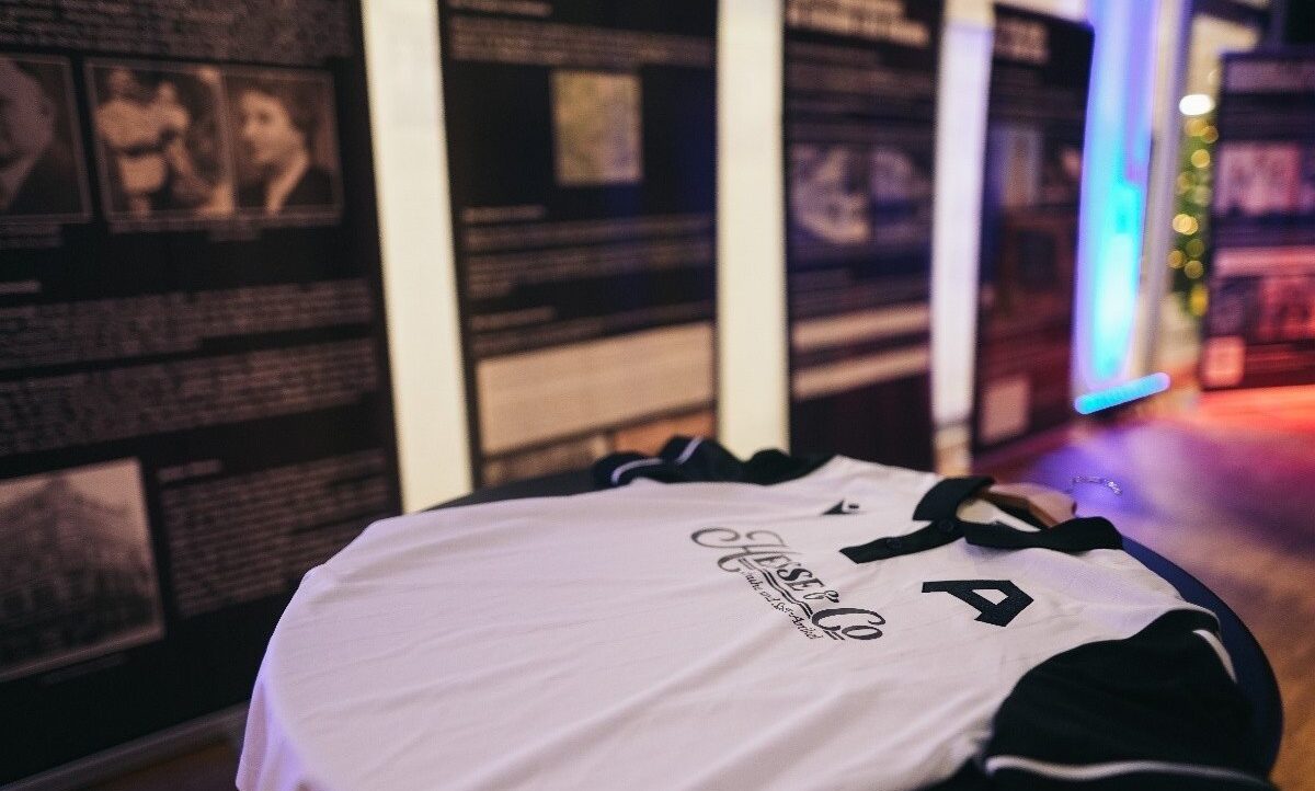 Ein weißes Trikot mit der Nummer 4 und schwarzem Text liegt auf einem Tisch. Im Hintergrund sind Bilder und Informationsplakate an einer Wand zu sehen.