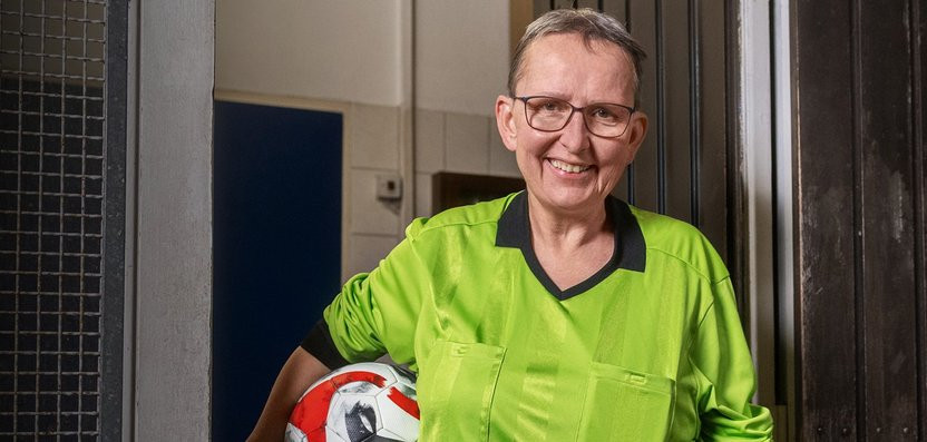 Eine lächelnde Frau in einem grünen T-Shirt steht in einem Flur und hält einen Fußball in ihren Armen. Sie trägt eine Brille und wirkt freundlich.