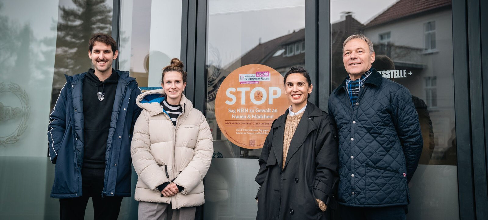 Vier Personen stehen vor einem Schaufenster, das ein Plakat mit der Aufschrift "STOP. Sag NEIN zu Gewalt an Frauen & Mädchen!" zeigt. Die Personen lächeln in die Kamera und tragen warme Jacken. Im Hintergrund sind Gebäude und Bäume zu sehen.