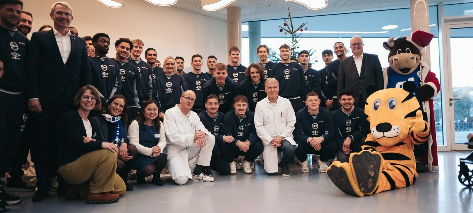Eine Gruppe von Menschen, darunter Spieler, Trainer und Mitarbeiter, posieren für ein Gruppenfoto in einem modernen Raum. Die Spieler tragen einheitliche Trainingskleidung mit dem Logo von Umbro und RB Leipzig. Zwei Maskottchen, ein Tiger und ein Bulle, sind ebenfalls Teil des Fotos. Im Hintergrund steht ein geschmückter Weihnachtsbaum.