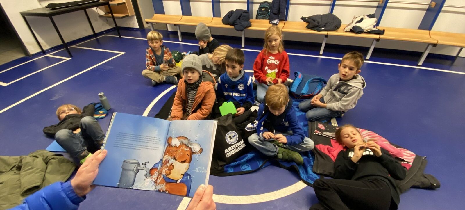 Eine Gruppe von Kindern sitzt im Kreis auf dem Boden einer Sporthalle und hört aufmerksam einer Person zu, die ihnen aus einem Buch mit einer Zeichnung eines Bären vorliest. Die Kinder tragen warme Kleidung und sitzen auf ihren Jacken und Taschen. Im Hintergrund sind eine Bank und ein Whiteboard mit einer Fußballtaktik zu sehen.