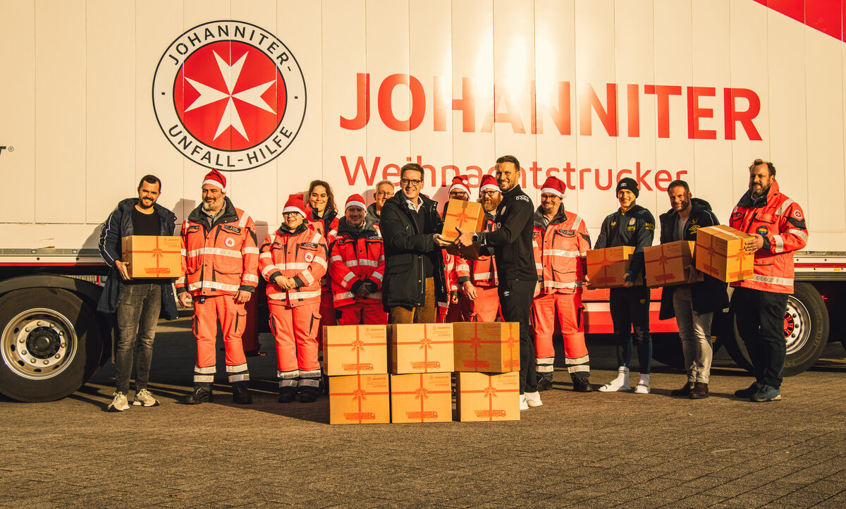 Eine Gruppe von Menschen, darunter Mitglieder der Johanniter-Unfall-Hilfe und Fußballspieler, stehen vor einem LKW mit dem Johanniter-Logo. Sie halten Kartons mit Weihnachtsgeschenken in den Händen und lächeln in die Kamera. Der LKW ist mit der Aufschrift "Johanniter Weihnachtstrucker" versehen.
