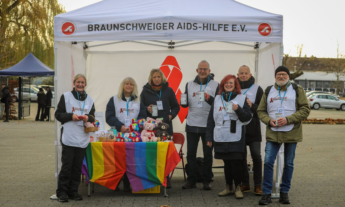 Eine Gruppe von sieben Personen steht vor einem Zelt mit der Aufschrift "BRAUNSCHWEIGER AIDS-HILFE E.V." Sie tragen weiße Westen mit Aufnähern und halten verschiedene Gegenstände in den Händen. Vor ihnen liegt ein Tisch mit einer Regenbogenflagge als Tischdecke, auf dem bunte Stofftiertiere und andere kleine Gegenstände liegen. Die Personen lächeln in die Kamera.